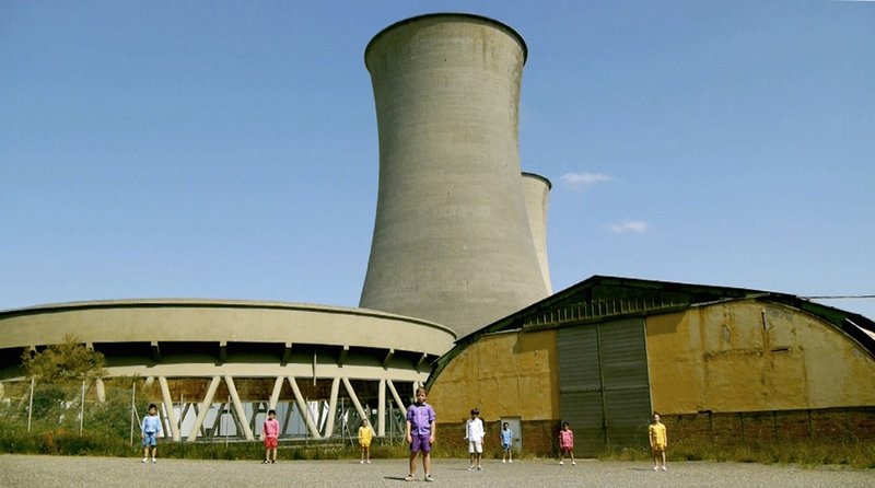 A still from Mikhail Karikis' film 'Children of Unquiet'. It shows children stood in front of a disused power plant.